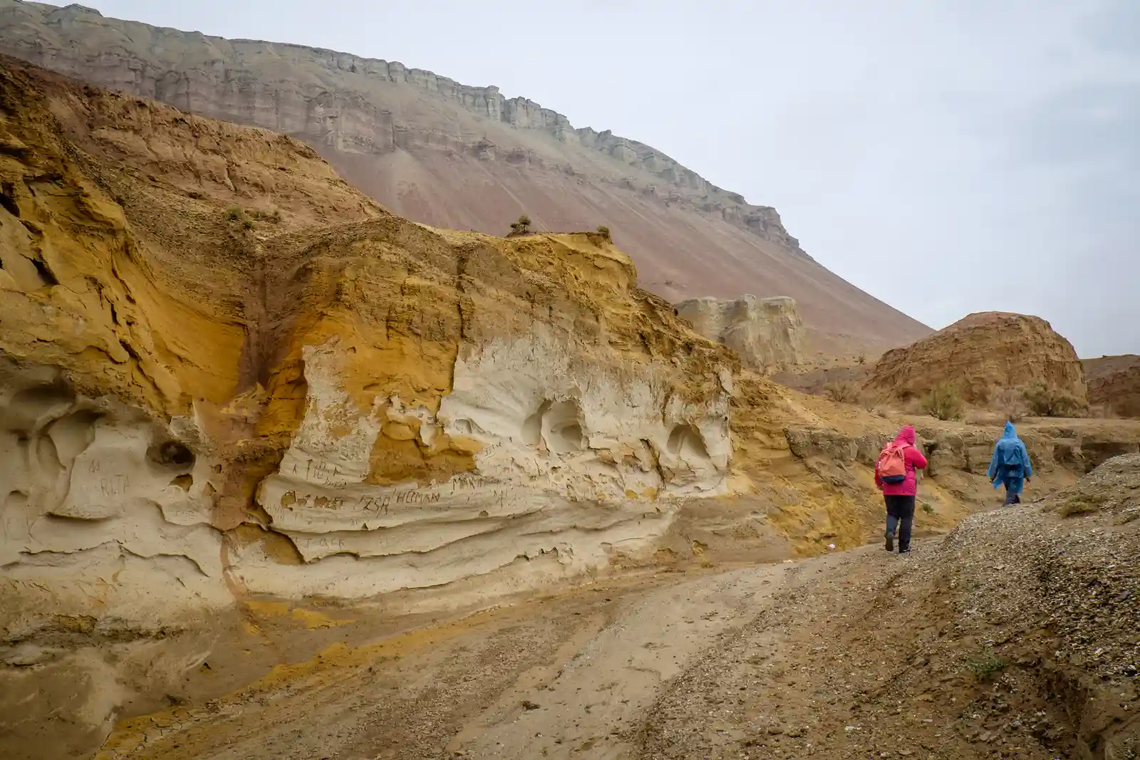 Two hikers in rain jackets walking a trail past eroded sandstone cliffs in the Aktau Mountains